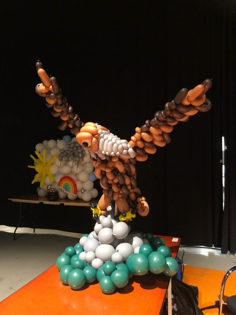 Een adelaar gemaakt van bruine, witte en oranje ballonnen staat op een sokkel van groene en grijze ballonnen op een oranje tafel. Op de achtergrond zijn nog meer ballonfiguren te zien.