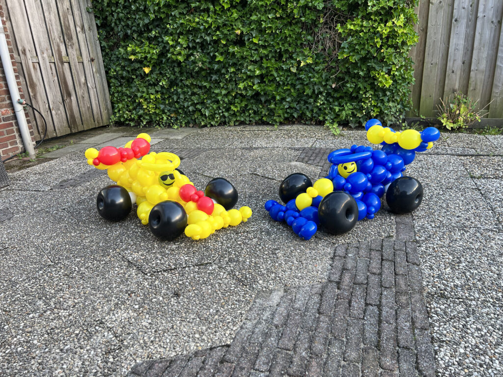 Twee autovormige ballonconstructies staan op een betegeld terras. De ene is geel met rood, de andere blauw met geel. Ze hebben zwarte ballonwielen.