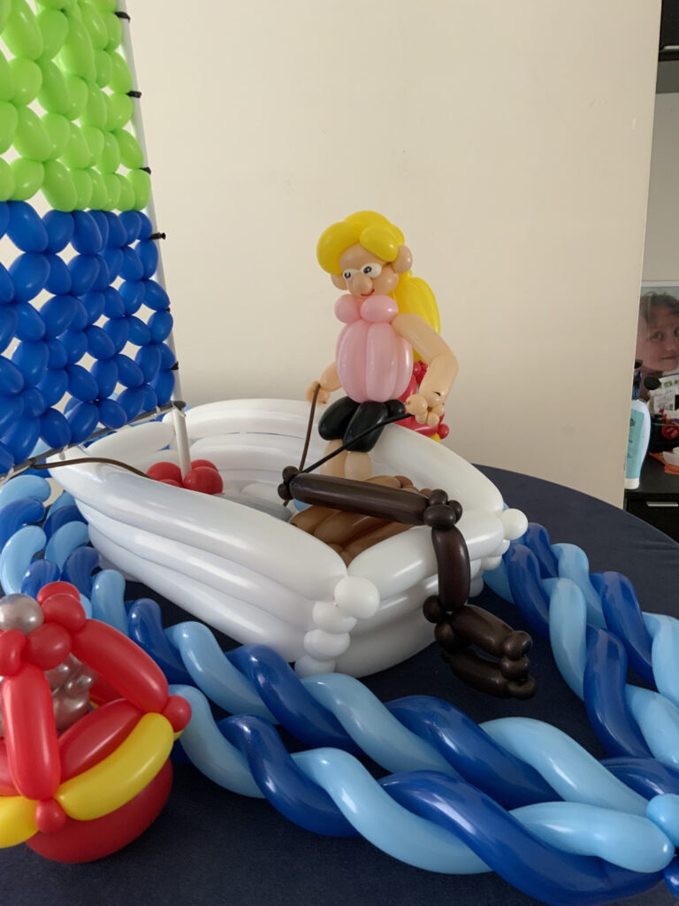 Een boot en poppetje gemaakt van ballonnen op een tafel. De boot is wit met bruin, het poppetje heeft roze en gele details, met blauwe en groene ballonzeilen.